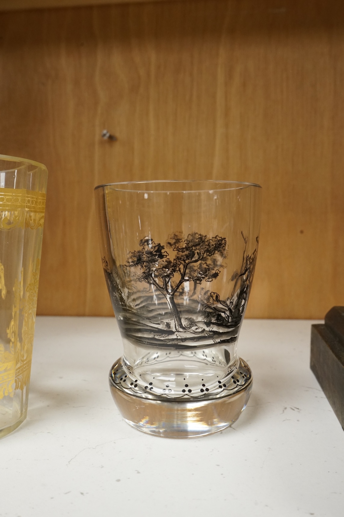 An Italian gilt-decorated and faceted beaker, inscribed Daniel Delvinis, the reverse with papal coat of arms, and a German Schwarzlot black enamelled ‘hunting’ beaker, largest 12cm (2)